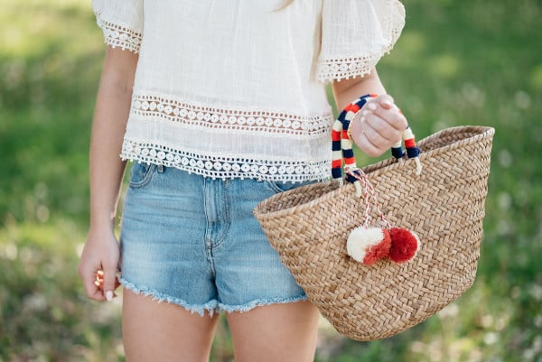 Bows & Sequins styles a pair of cut off shorts with a gauzy off-the-shoulder top, Jack Rogers sandals, and a Kayu pom pom tote.