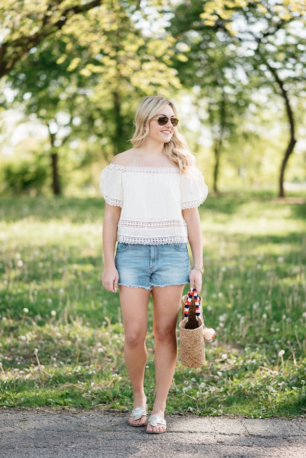 Bows & Sequins styles a pair of cut off shorts with a gauzy off-the-shoulder top, Jack Rogers sandals, and a Kayu pom pom tote.