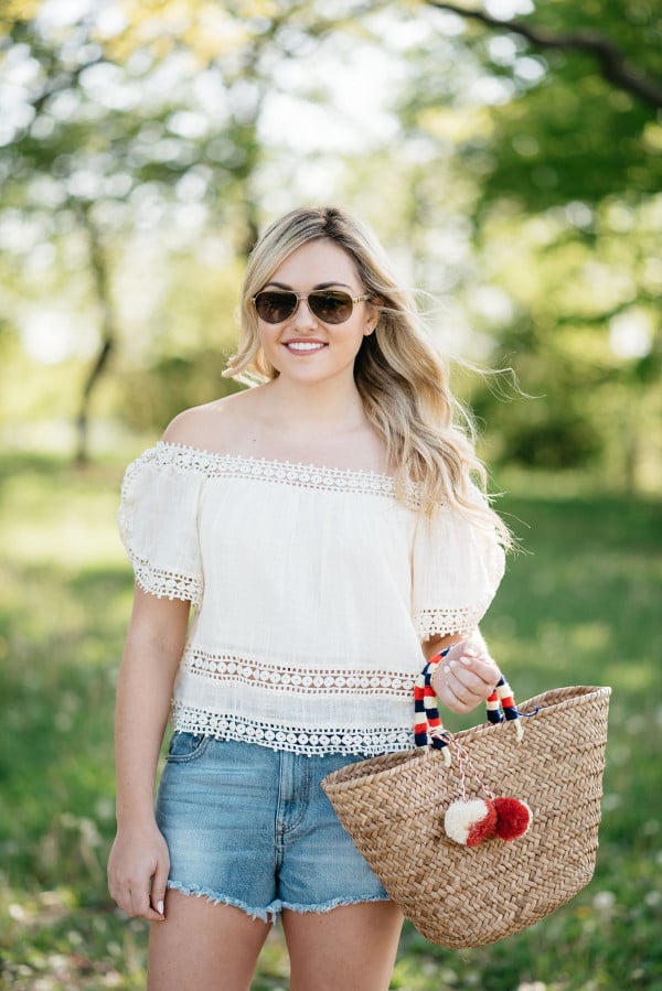 Bows & Sequins styles a pair of cut off shorts with a gauzy off-the-shoulder top, Jack Rogers sandals, and a Kayu pom pom tote.