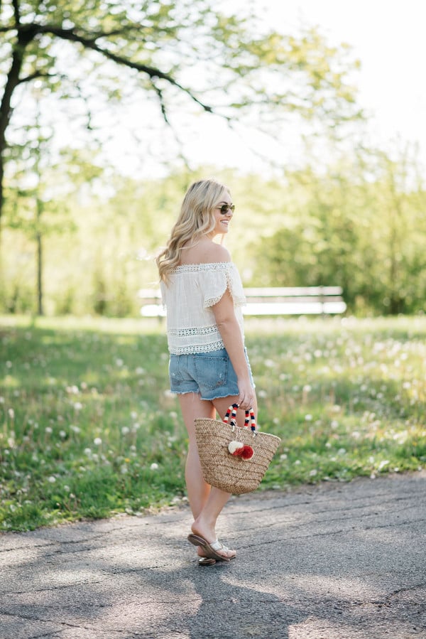 Bows & Sequins styles a pair of cut off shorts with a gauzy off-the-shoulder top, Jack Rogers sandals, and a Kayu pom pom tote.