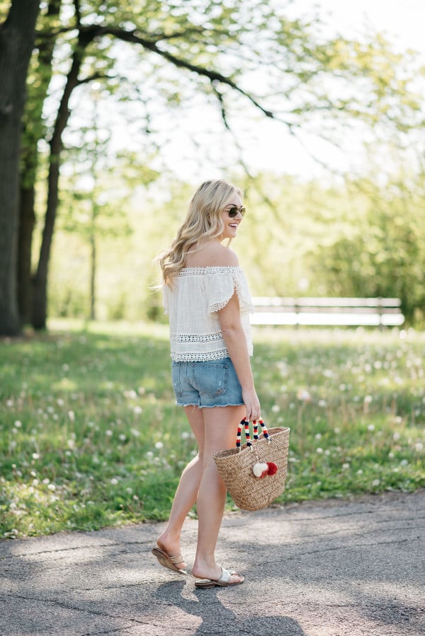 Bows & Sequins styles a pair of cut off shorts with a gauzy off-the-shoulder top, Jack Rogers sandals, and a Kayu pom pom tote.