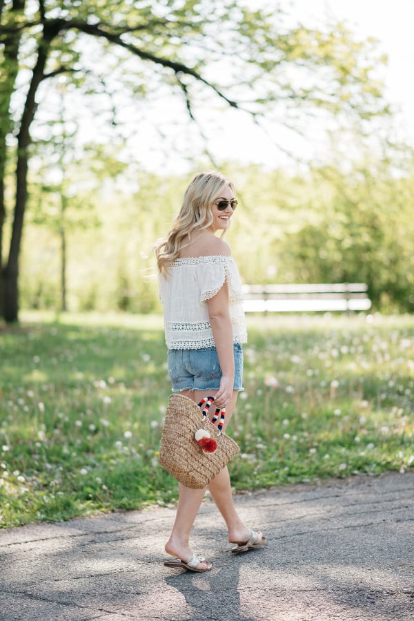 Bows & Sequins styles a pair of cut off shorts with a gauzy off-the-shoulder top, Jack Rogers sandals, and a Kayu pom pom tote.