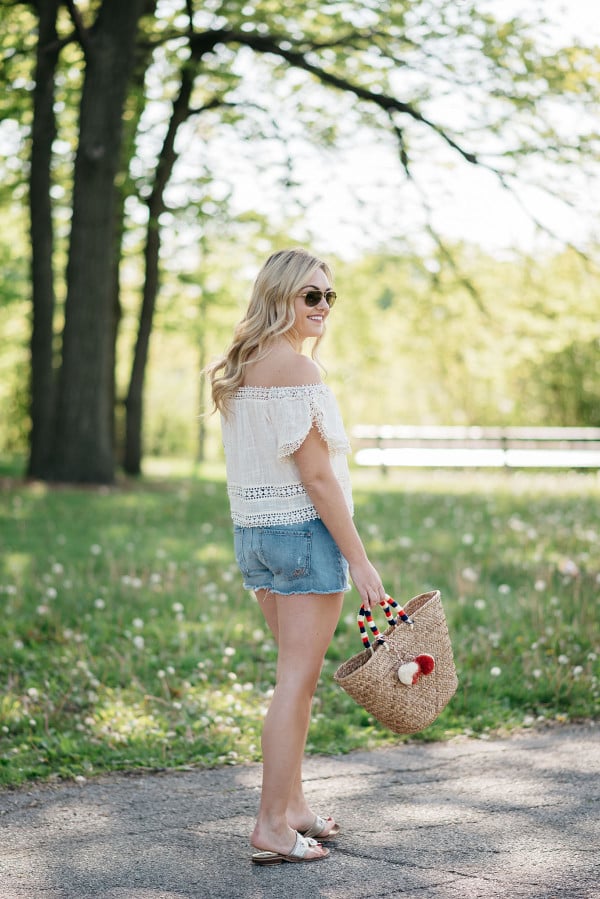 Bows & Sequins styles a pair of cut off shorts with a gauzy off-the-shoulder top, Jack Rogers sandals, and a Kayu pom pom tote.