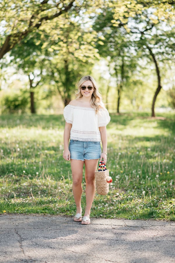 Bows & Sequins styles a pair of cut off shorts with a gauzy off-the-shoulder top, Jack Rogers sandals, and a Kayu pom pom tote.