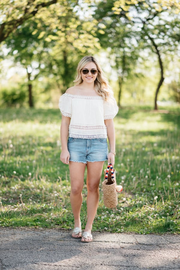 Bows & Sequins styles a pair of cut off shorts with a gauzy off-the-shoulder top, Jack Rogers sandals, and a Kayu pom pom tote.