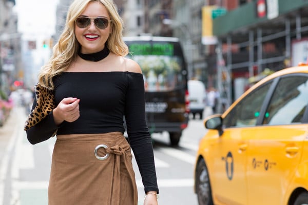 Bows & Sequins wearing a black off-the-shoulder crop top, a black choker, a suede wrap skirt with a thigh slit, and lace-up black heels in New York City by the iconic Flatiron Building near Madison Square Park.