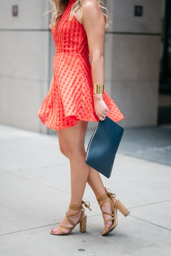 Bows & Sequins wearing a Lovers + Friends red-orange dress with a gold cuff, a navy blue leather clutch, and nude lace-up suede sandals.