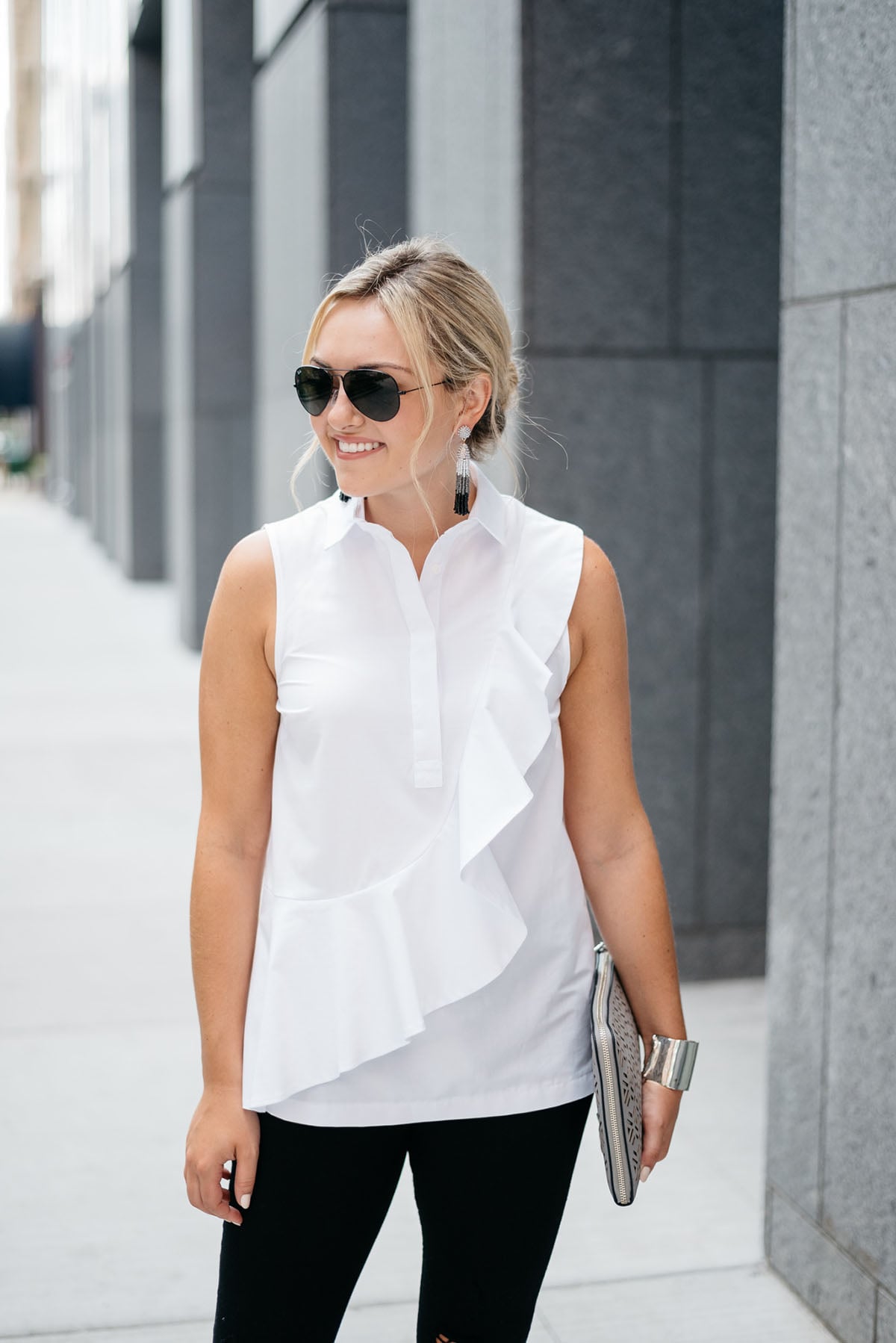 Bows & Sequins styling a white ruffled sleeveless collared shirt, black destroyed denim, tassel drop earrings, a metallic cut-out clutch, and black Ray-Ban aviator sunglasses.