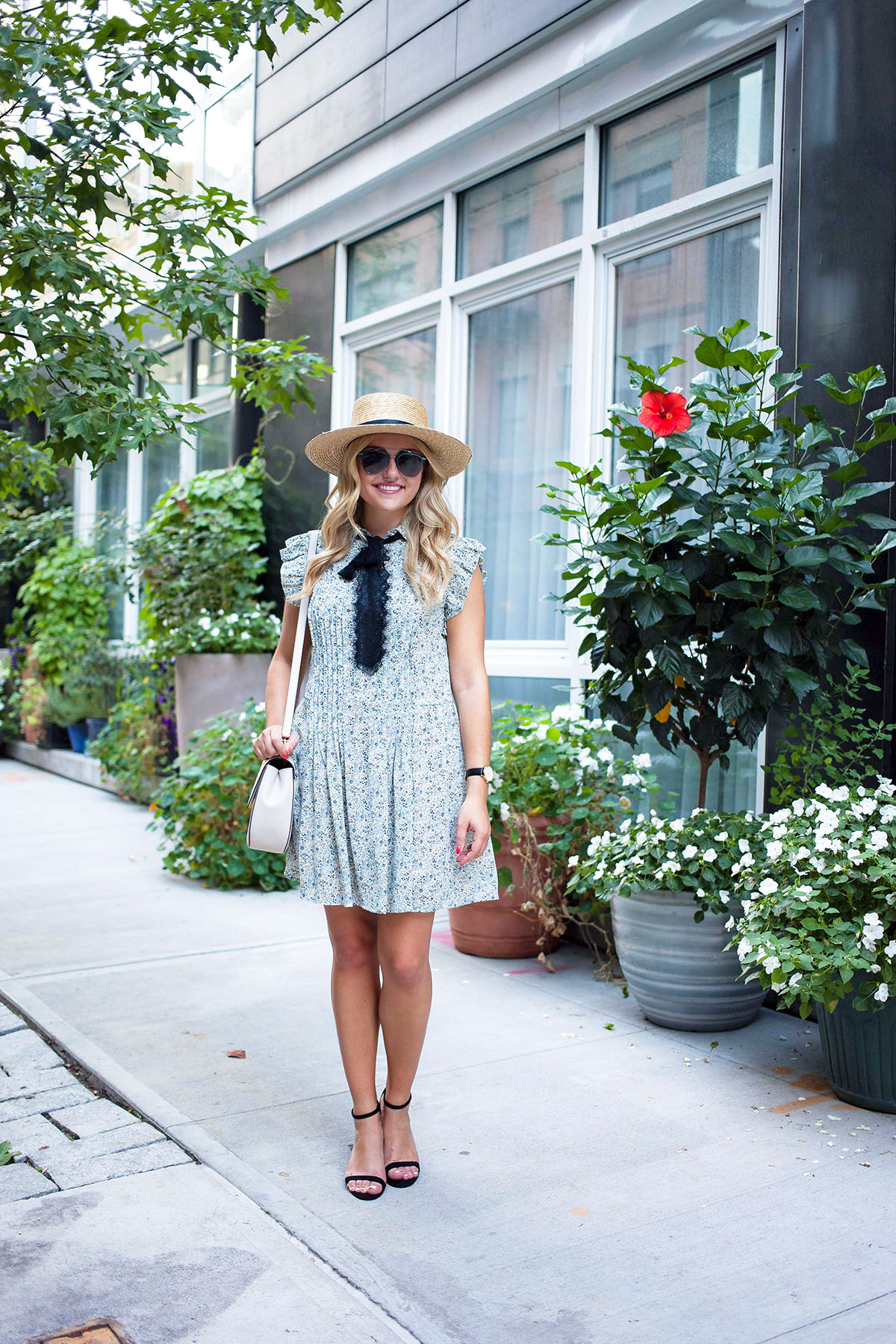 Fashion blogger Jessica Sturdy of Bows & Sequins styling a blue floral dress with a bow, straw hat, sunglasses, and Kate Spade purse in New York City.