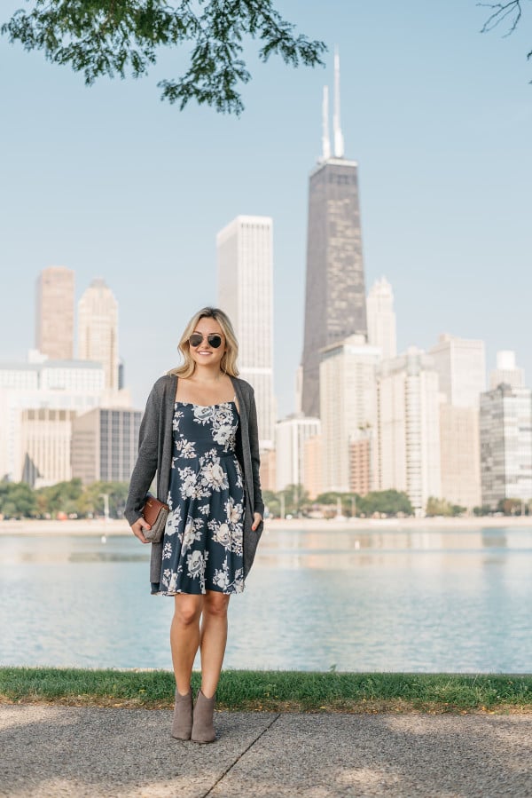 Jessica Sturdy of Bows & Sequins stands in front of Chicago wearing a navy floral dress, long grey sweater, and aviator sunglasses.