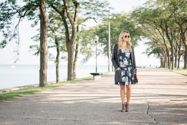 Fashion blogger Bows & Sequins wears an outfit consisting of a navy floral cami dress, long grey sweater, booties, aviator sunglasses, and a clutch purse.