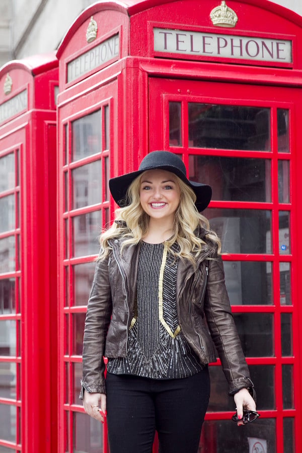 Jessica Sturdy of Bows & Sequins, a fashion-focused lifestyle and travel blog, styling a moto jacket, black hat, and a beaded tank in front of a phone booth in London. 