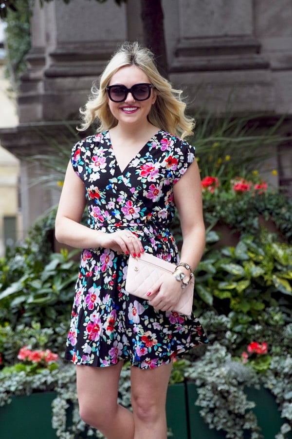 Travel and fashion blogger Bows & Sequins styling a navy floral wrap dress, sunglasses, and a blush leather clutch purse in London.