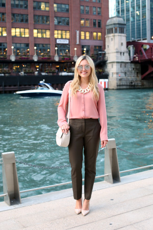 Bows & Sequins wearing a cute fall work outfit. She paired a silky pink blouse with olive green slim-fit ankle trousers, nude pointed toe pumps, a cream-colored crossbody bag, a white link statement necklace, and blush pink sunglasses.
