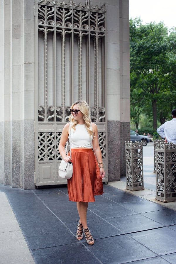 Pleated skirts are one of fall's hottest trends! Blogger Bows & Sequins styles hers with a crop-top tank, lace-up leopard heels, oversized sunglasses, and a round crossbody bag.
