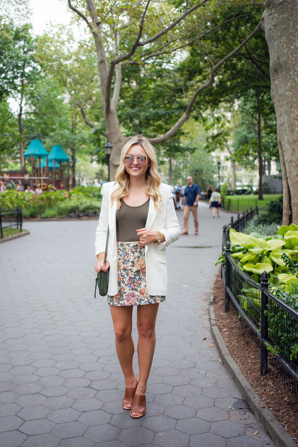 Bows & Sequins wearing a Free People olive green body suit, a Zara floral mini skirt, a white Express Edition blazer, and J.Crew leather mules for a cute summer/fall outfit. Accessorized with a green crossbody bag and blush pink mirrored sunglasses.