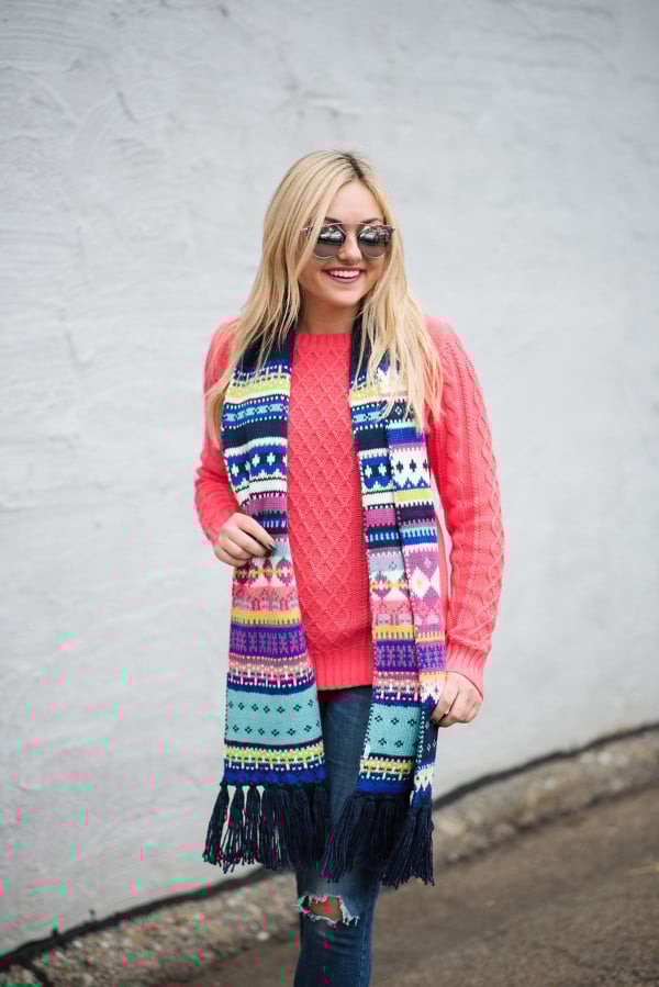 Bows & Sequins wearing a neon pink sweater with a fair isle scarf, blue jeans, and silver mirrored sunglasses.