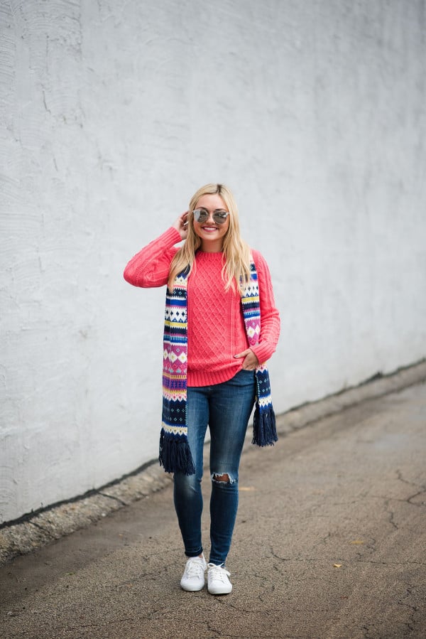 Bows & Sequins wearing a neon pink sweater with a fair isle scarf, blue jeans, silver mirrored sunglasses, and white sneakers