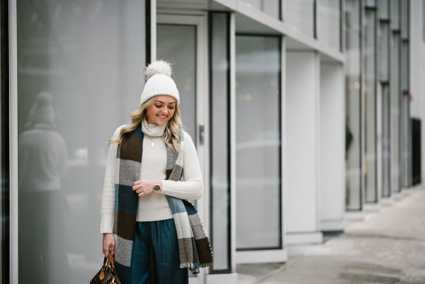 Bows & Sequins wearing a turtleneck sweater, a fur pom pom beanie, a checkered blanket scarf, and chambray wide leg pants.