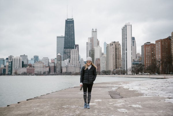 Bows & Sequins wearing a black puffer jacket and a grey beanie in front on the Chicago skyline at North Avenue Beach.