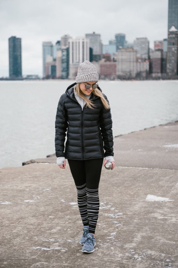 Bows & Sequins wearing a black puffer jacket and striped black and white leggings in front of the Chicago skyline by Lake Michigan.