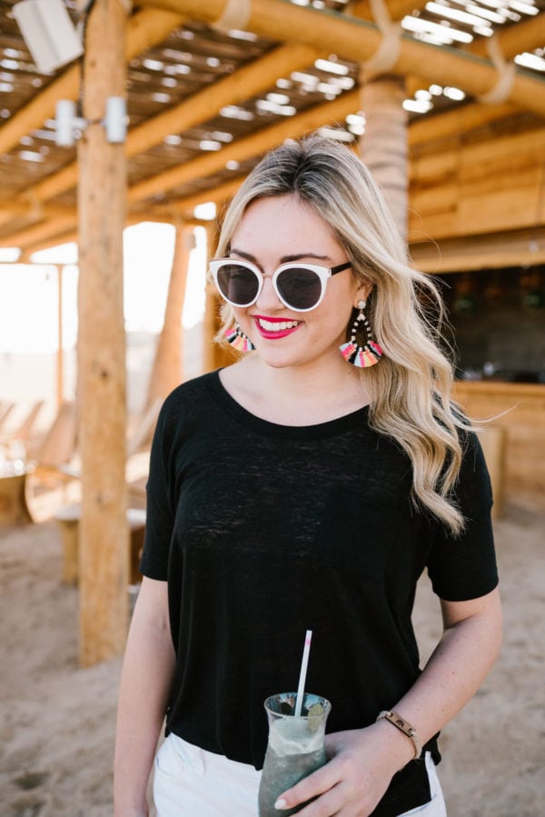 Bows & Sequins wearing striped colorful fringe Baublebar earrings with a black tee and black and white sunglasses in Cabo.