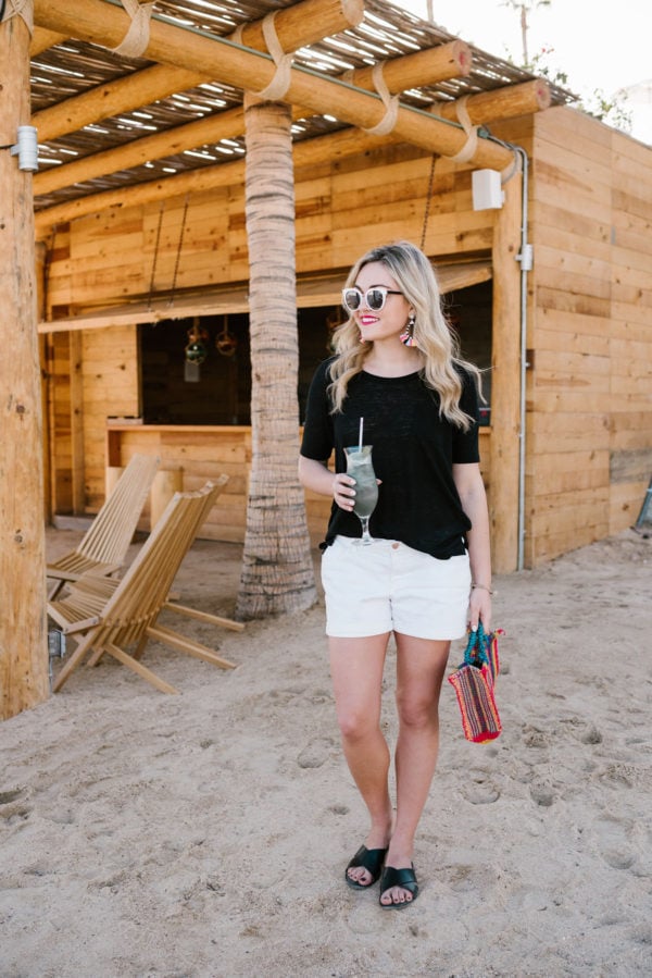 Travel blogger Bows & Sequins wearing a black tee and white shorts with colorful accessories by a tiki hut in Cabo.