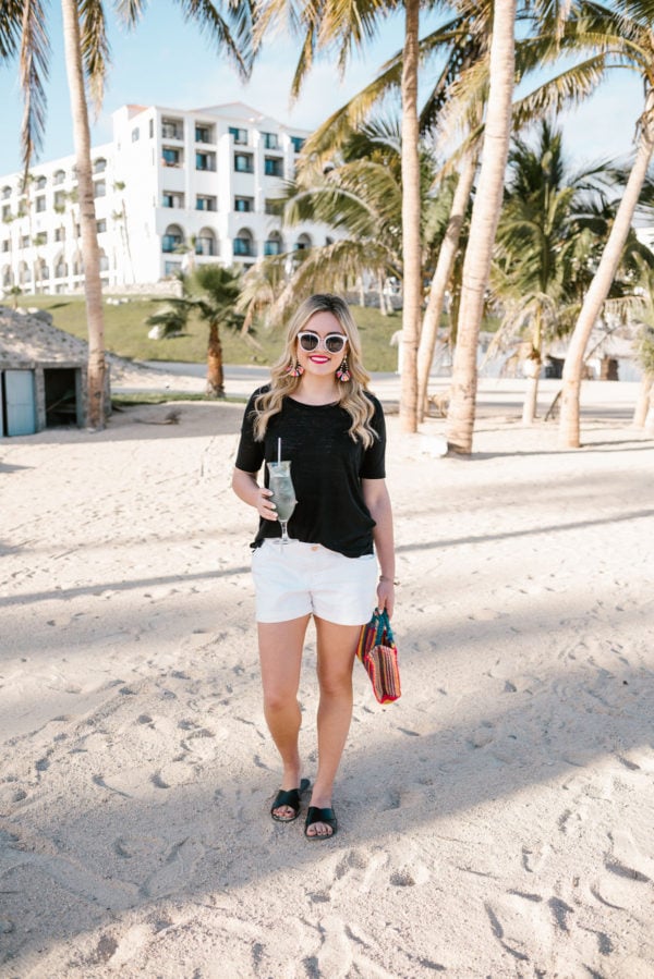 Bows & Sequins wearing a black boyfriend tee and white Old Navy shorts on the beach in Cabo with palm trees.
