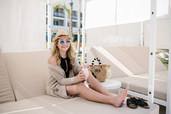 Bows & Sequins wearing a black one-piece swimsuit and a pom pom hat in a cabana in Cabo San Lucas Mexico.