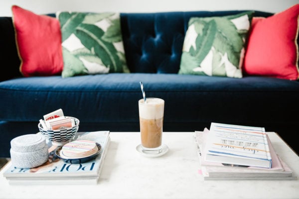 Bows & Sequins sharing a recipe to make iced almond lattes at home. A glass of iced coffee sitting on a marble coffee table with stacks of magazines and coffee table books, with a navy velvet tufted sofa and palm leaf pillows in the background.