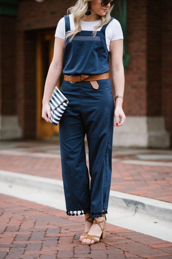Bows & Sequins wearing a navy blue jumpsuit with tassels, a leather belt, lace-up sandals, and a striped clutch.