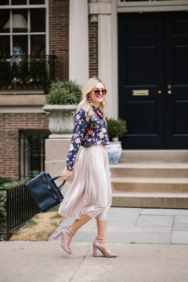 Bows & Sequins wearing a pink pleated midi skirt and floral blouse with a navy handbag and Kate Spade pumps.