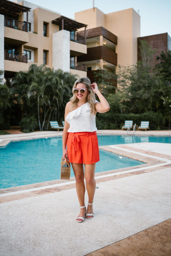 Bows & Sequins wearing a single shoulder ruffle top from Old Navy, orange tie-waist shorts, and J.Crew pom pom sandals.