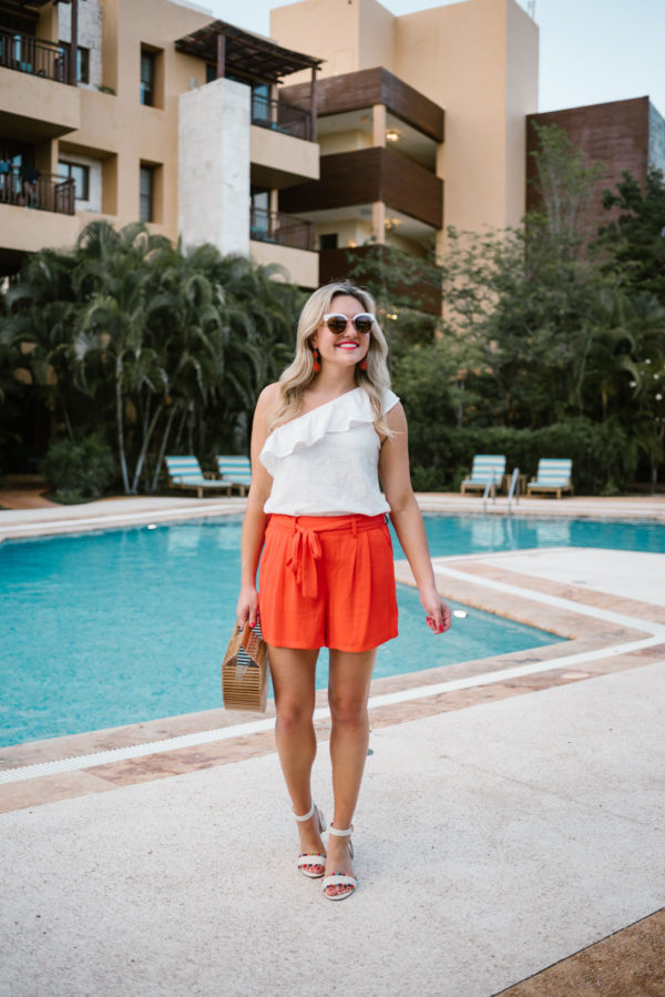 Bows & Sequins wearing a one-shoulder ruffle top and tie-waist shorts from Old Navy with mirrored sunglasses and J.Crew pom-pom sandals.