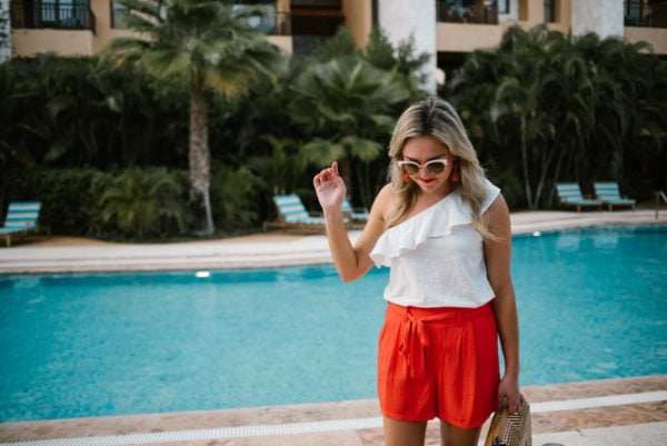 Bows & Sequins wearing a one-shoulder ruffle top with white sunglasses and orange Old Navy shorts.