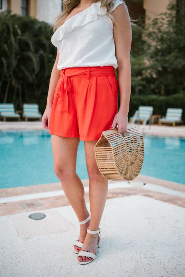 Jessica of Bows & Sequins, a Chicago based travel and lifestyle blog, wearing orange Old Navy tie-waist shorts with J.Crew pom-pom sandals and a bamboo handbag.