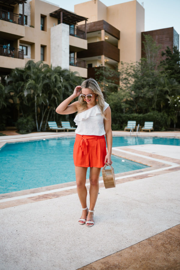 Travel and lifestyle blogger Bows & Sequins wearing a white Old Navy ruffle top with orange tie-waist shorts and a Cult Gaia Ark bamboo handbag.