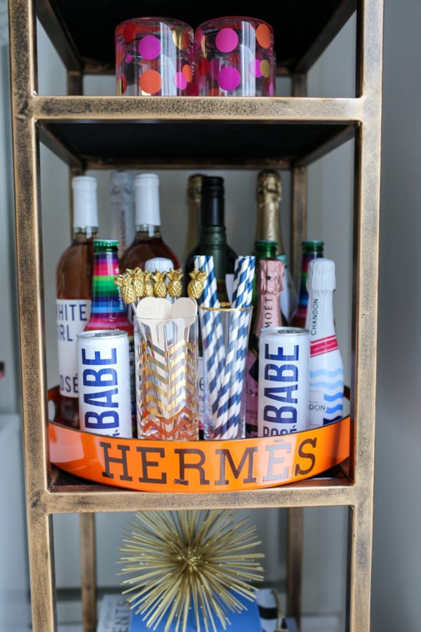 Bows & Sequins shares her bar cart staples on an Hermès orange tray: White Girl rosé, Moët, striped straws, pineapple drink stirrers.