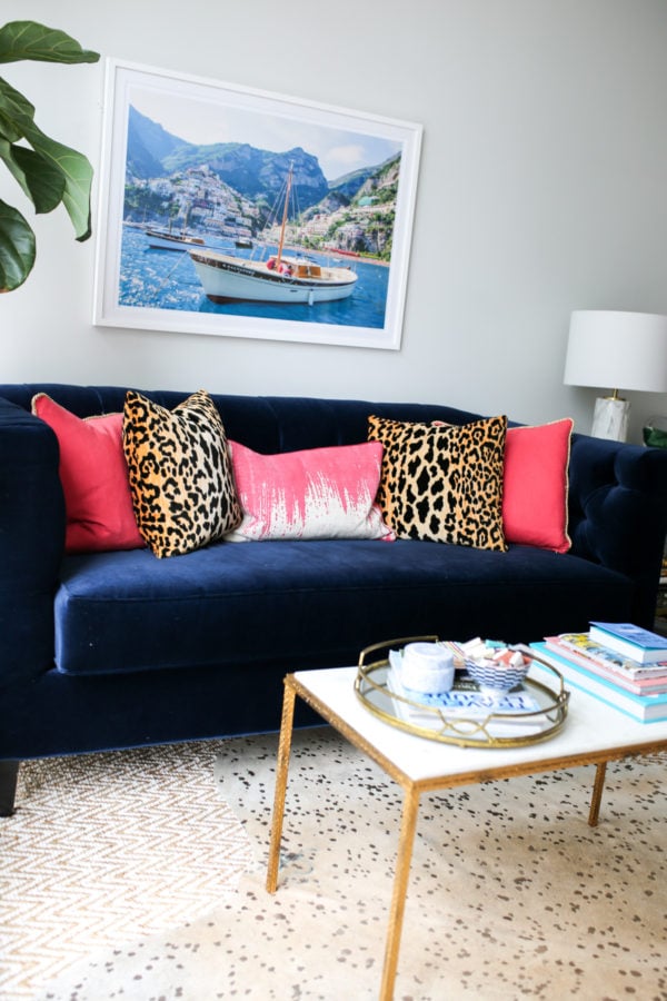 Bows & Sequins navy velvet Society Social couch with pink and leopard throw pillows, framed Gray Malin Italy print, and gold and marble coffee table with mirror tray.
