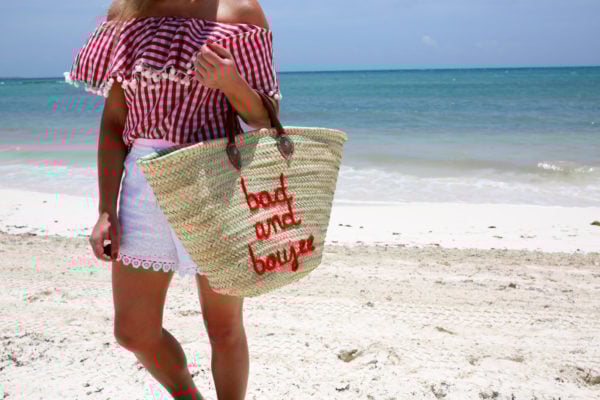 Bows & Sequins wearing a gingham pom pom hem off the shoulder top and white eyelet shorts with a Bad and Boujee straw tote in Mexico.