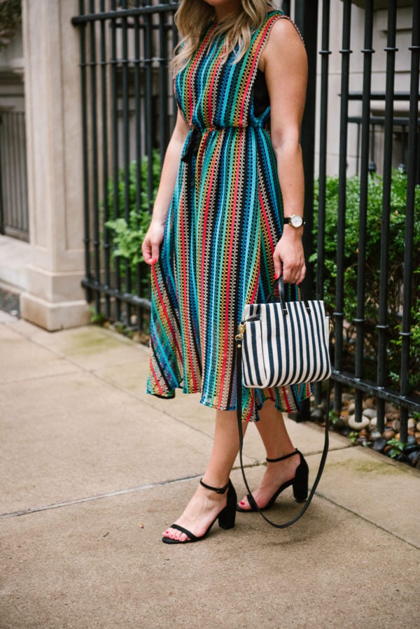 Jessica of Bows & Sequins, a fashion-focused lifestyle blog, wearing a rainbow striped crochet dress, a navy striped bag, and black sandals.
