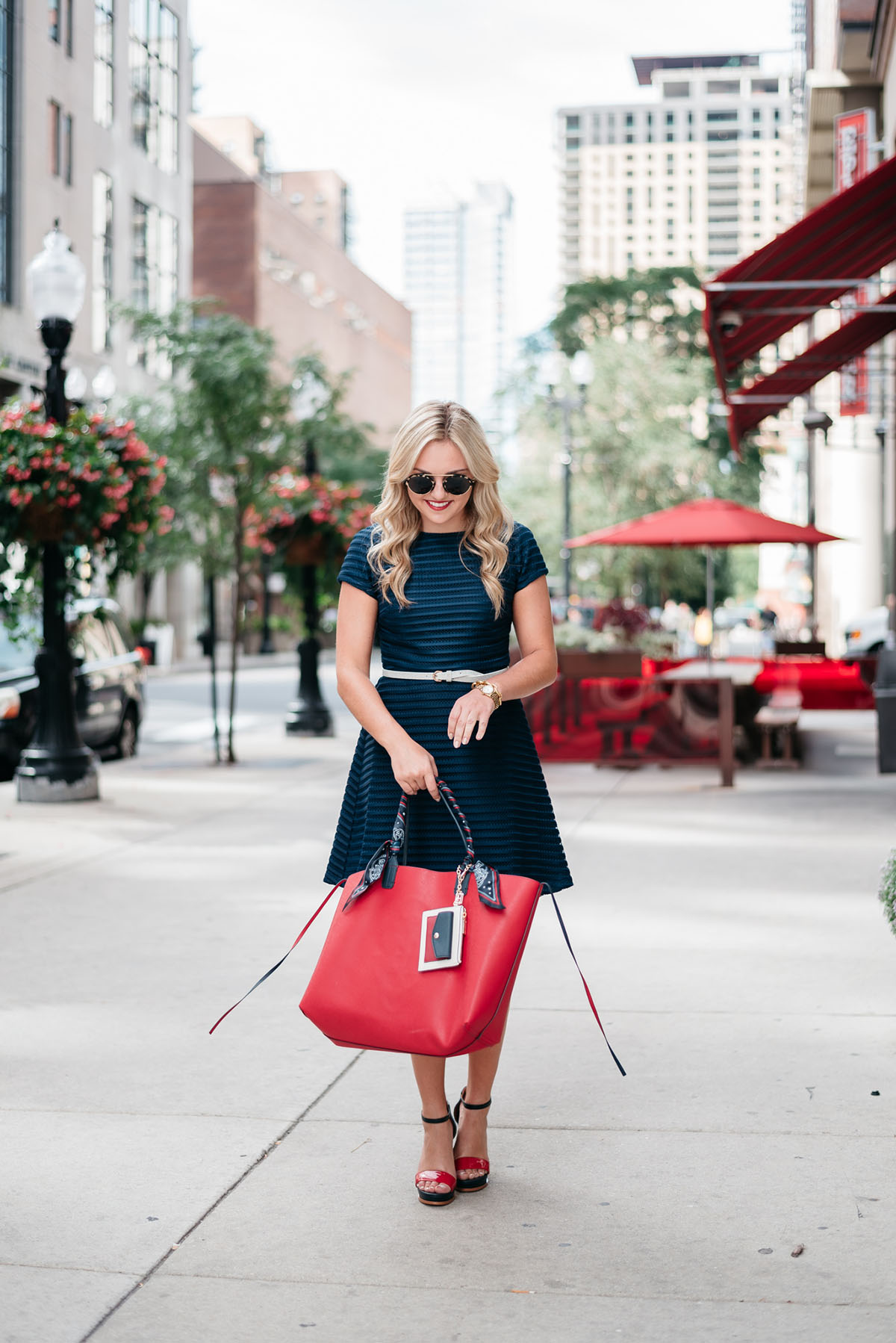 Bows & Sequins holding a Gigi x Tommy collaboration red reversible tote and wearing a navy fit and flare dress.