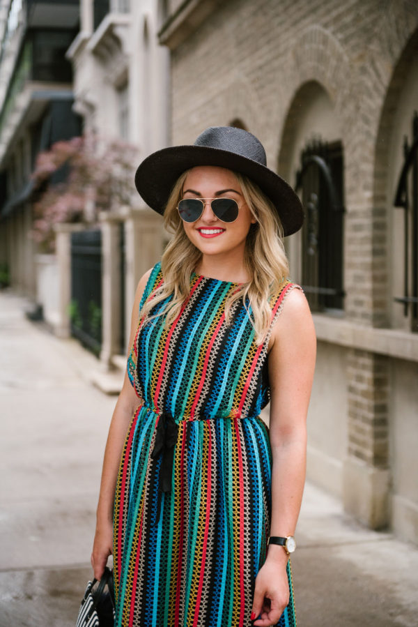 Fashion-focused lifestyle blogger Bows & Sequins wearing a rainbow striped sleeveless dress with a black straw hat and gold aviators.