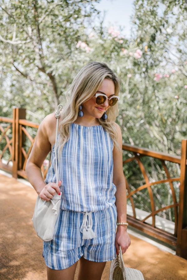 Bows & Sequins wearing white rim sunglasses, blue statement earrings, a blue and white striped romper, and a Clare V bucket bag.