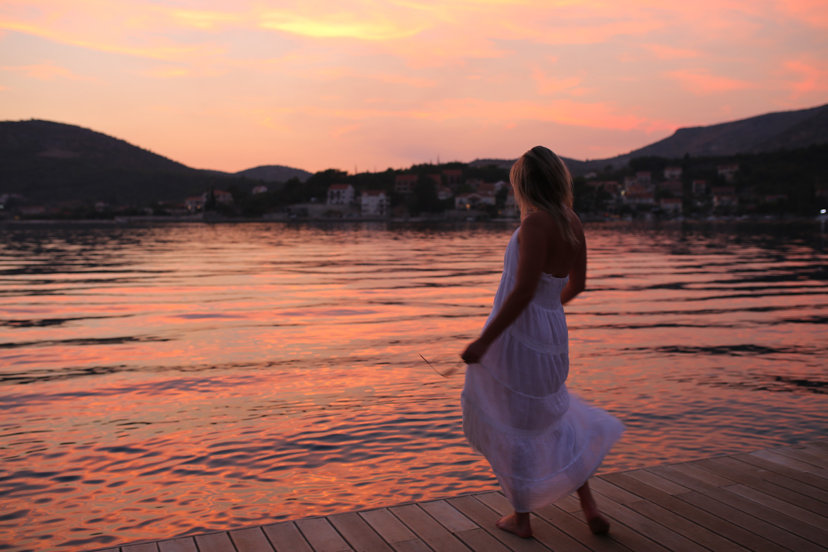 Travel blogger Bows & Sequins wearing a white maxi dress on a dock in Croatia at sunset.