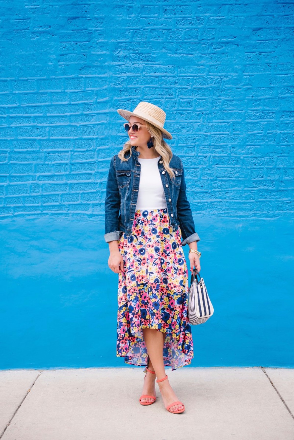 Chicago-based lifestyle blogger Bows & Sequins styling a high-low floral dress with coral sandals, a straw hat, and a dark denim jacket.