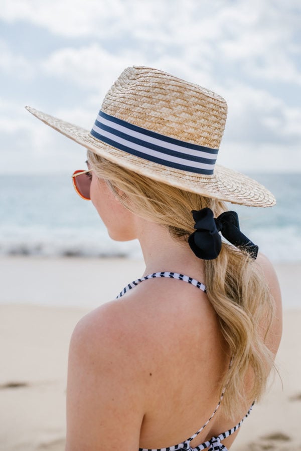 Bows & Sequins wearing a striped straw hat with a hair bow and a swimsuit on the beach.