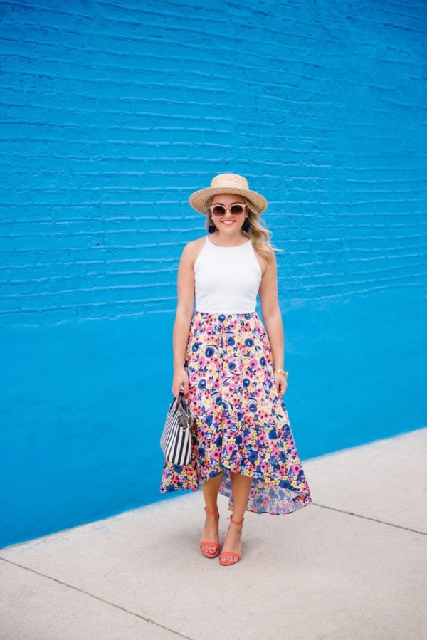 Jessica of Bows & Sequins, a fashion-focused lifestyle blog, wearing a floral Hutch dress from Anthropologie, a straw hat, Old Navy coral heels, Nordstrom sunglasses, a Julie Vos bracelet, and BaubleBar earrings with a Clare V striped tote.