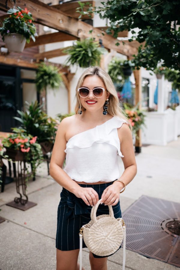 Jessica of Bows & Sequins, a fashion-focused lifestyle blog, wearing an ASTR ruffle one-shoulder crop top and a J.O.A. pinsstripe skirt with Call It Spring sunglasses and a Clare V straw bag.
