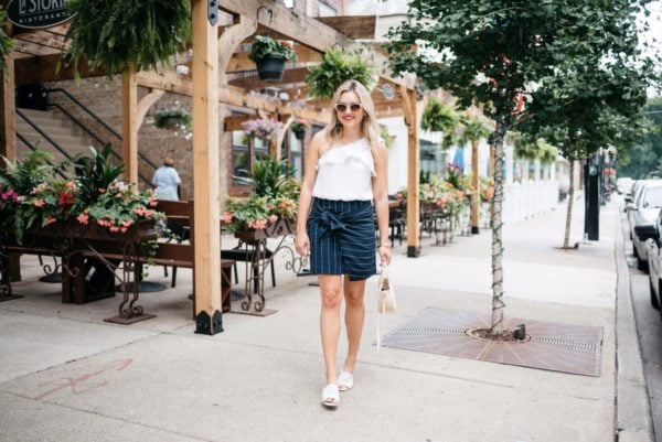 Bows & Sequins wearing a ruffle one-shoulder crop top and a navy pinstripe tie-front skirt with Call It Spring sunglasses and Kurt Geiger slides.